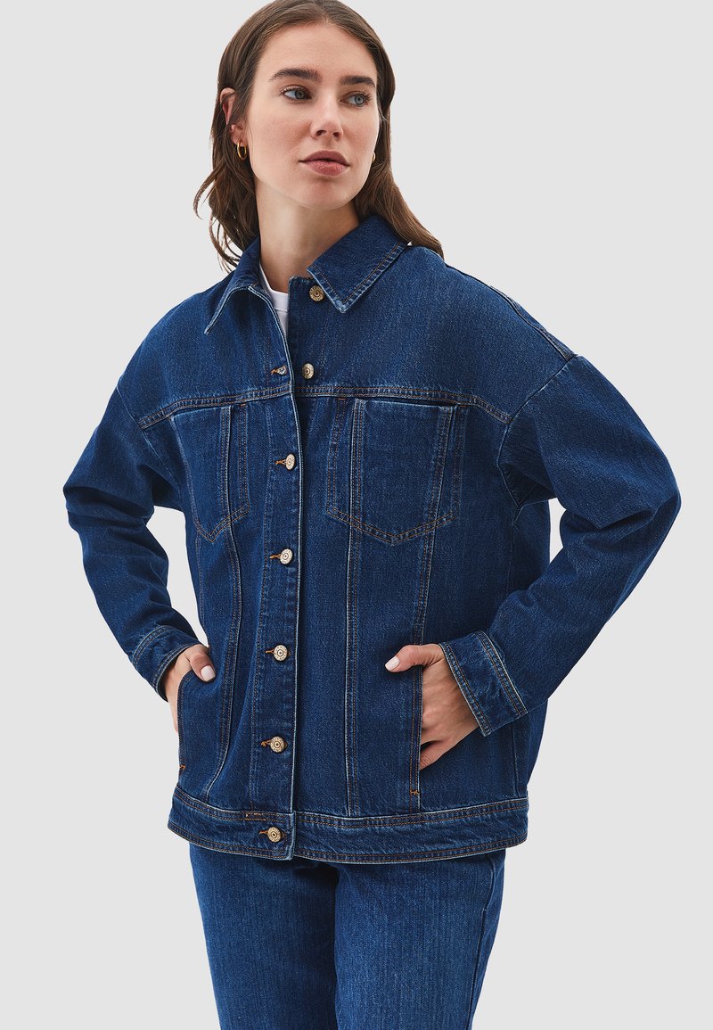 Kvinna i mörkblå denimjacka och jeans, med händerna i jackfickorna, tittar åt sidan mot en enkel grå bakgrund.