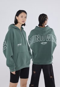 Πράσινη oversized hoodie με τσέπη καγκουρό, διαθέτει λευκό γραφικό κείμενο "SOUNDWAVE" στην πλάτη και λογότυπο μπροστά. Φοριούνται μαύρες βερμούδες.