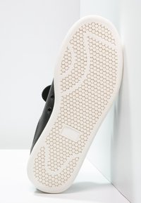 Svart sko med en texturerad gummisula, prydd med cirkulära mönster. Sulan är vit med intrikata greppdetaljer och en rektangulär accent.