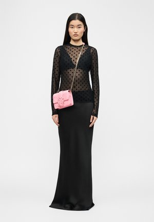 Frau trägt ein schwarzes, durchsichtiges Oberteil mit Herzmuster, einen langen schwarzen Rock und eine kleine pinke Umhängetasche mit goldener Kettenriemen.