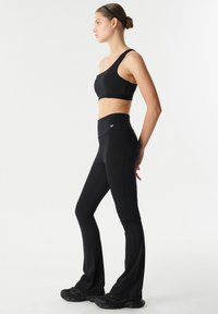 Vrouw die zijwaarts staat in een zwarte sportbeha met één schouder, hoge zwarte leggings en zwarte sneakers tegen een effen witte achtergrond.