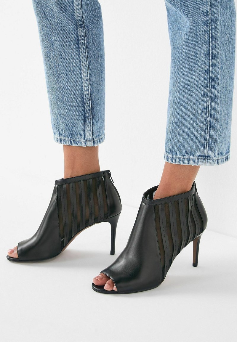 Bottines noires à bout ouvert avec panneaux transparents rayés et talons aiguilles, portées avec un jean court en denim bleu clair sur fond blanc.