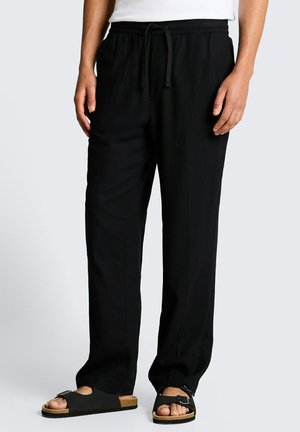 Person trägt lose schwarze Kordelzughose und schwarze offene Sandalen mit Schnallen, steht vor einem schlichten hellen Hintergrund.