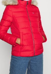 Chaqueta acolchada roja con capucha de piel sintética, con cierre de cremallera, acolchado horizontal y bolsillos laterales. Combinada con vaqueros azules.