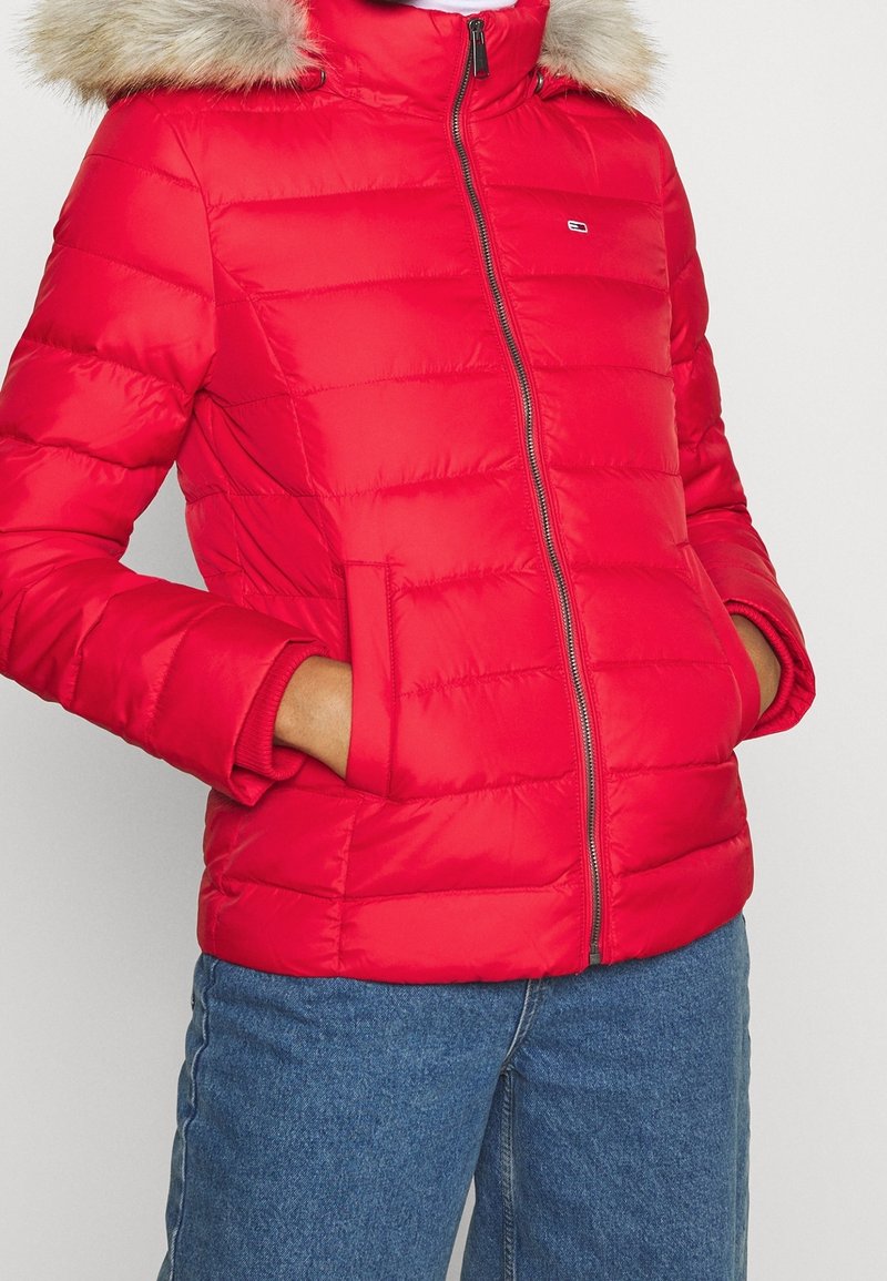 Chaqueta acolchada roja con capucha de piel sintética, con cierre de cremallera, acolchado horizontal y bolsillos laterales. Combinada con vaqueros azules.