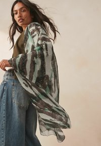 Sheer kimono in green and black abstract pattern with ribbed texture and embellished accents. Lightweight fabric drapes loosely.