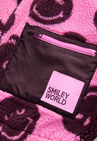 Tissu polaire rose et violet foncé avec motif de visages souriants, comprenant une poche zippée noire avec un patch rose portant l’inscription « SMILEY WORLD ».