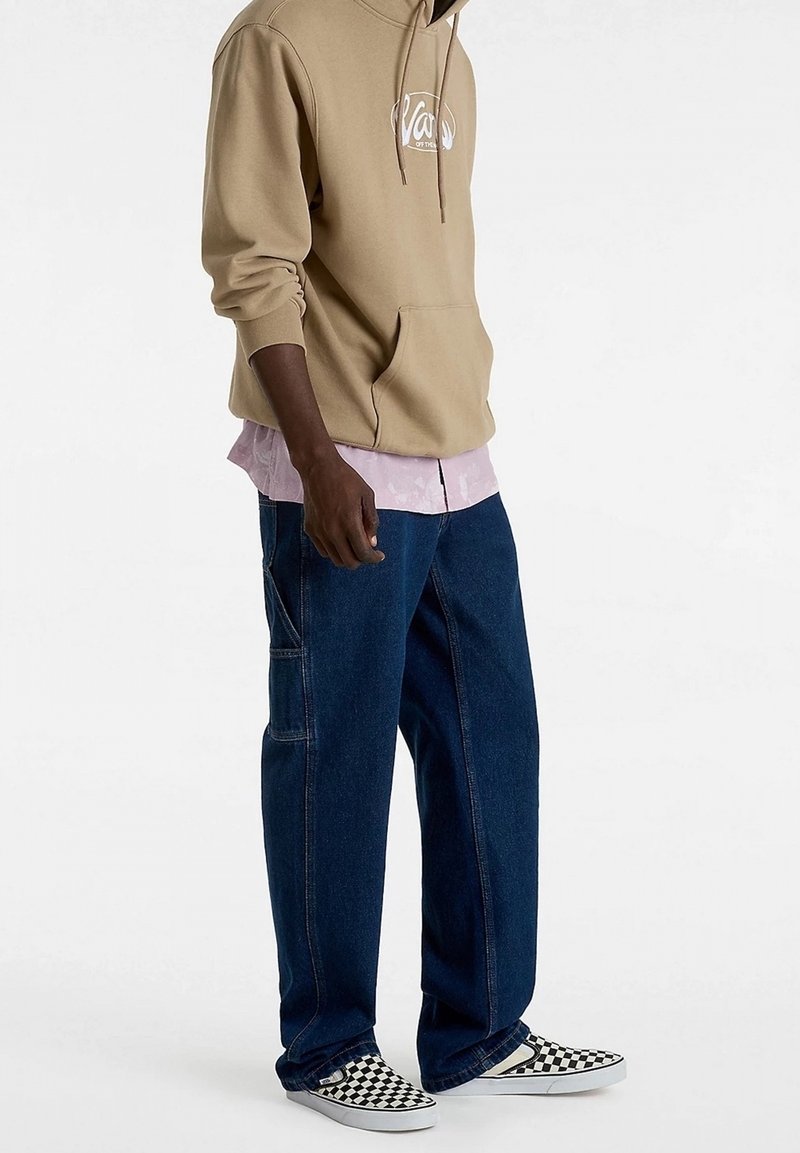 Personne portant un sweat à capuche beige, une chemise rose, un jean bleu foncé ample et des chaussures slip-on à carreaux noirs et blancs, debout sur un fond blanc uni.