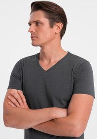 Muž se založenýma rukama, oblečený v tmavě šedém tričku s výstřihem do V, dívající se vlevo na pozadí čistě bílé.