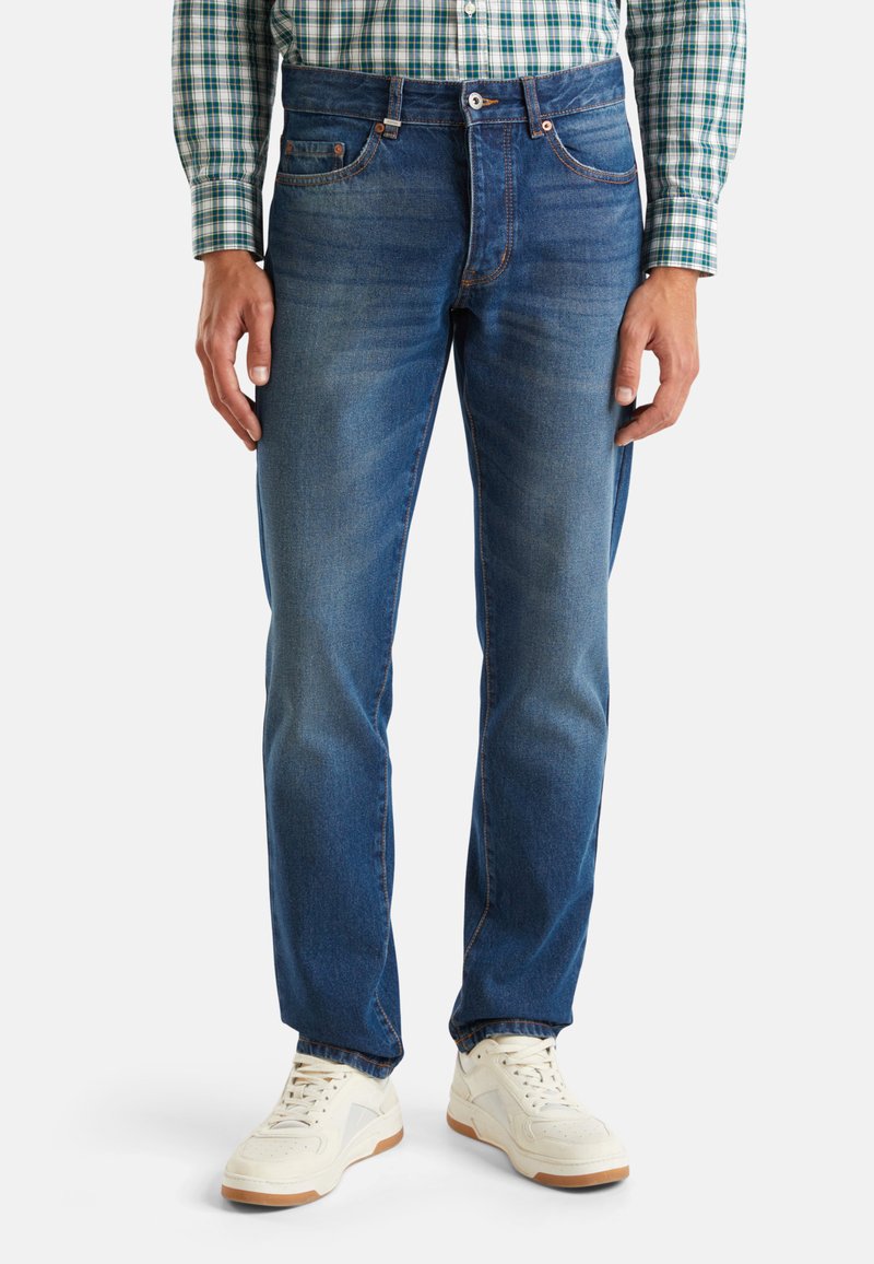 Donkerblauwe denim jeans met een rechte pijp, voorzien van voorzakken, een ritssluiting en subtiele vervaging voor textuur. Gecombineerd met witte sneakers.
