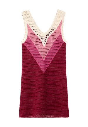 Femme debout contre un mur blanc portant une robe sans manches en crochet dans des tons de rose et de bordeaux avec des tongs dorées et noires.
