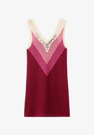 Femme debout contre un mur blanc portant une robe sans manches en crochet dans des tons de rose et de bordeaux avec des tongs dorées et noires.