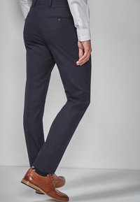 Marineblaue Anzughose mit schlanker Passform, hergestellt aus glattem Stoff, mit hinteren Taschen und Knopfverschlüssen, kombiniert mit braunen Schuhen.