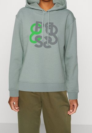 Osoba w jasnozielonej bluzie z kapturem z abstrakcyjnymi literami w odcieniach zieleni i szarości, w połączeniu z oliwkowozielonymi spodniami.