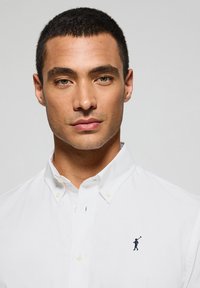 Wit overhemd met knoopsluiting en een puntige kraag, voorzien van een klein marineblauw logo van een golfer op de linkerborst, gemaakt van gestructureerde stof.