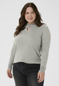 Vrouw met lang bruin haar, gekleed in een lichtgrijze lange mouwtop met een kleine keyhole uitsnijding bij de kraag en zwarte broek, met een lichte glimlach.