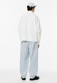 Camicia bianca oversize con spalle scese abbinata a jeans larghi di colore blu chiaro; il modello indossa un cappellino grigio chiaro e stivaletti neri.