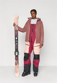 Kvinna iklädd en rosa och vinröd skidjacka och skidbyxor, som håller en ensam rosa och svart skid vertikalt framför en neutral bakgrund.