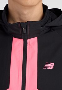 Veste noire avec accents roses, dotée d'un col haut, d'une fermeture éclair intégrale et du logo New Balance. Texture lisse avec des coutures contrastées.