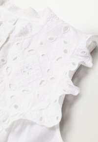 Top blanco de algodón sin mangas que presenta recortes bordados, un torso texturizado y un borde con forma de ondas en los hombros para mayor detalle.