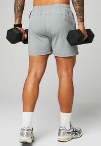 Graue Sportshorts mit elastischem Bund, hält schwarze Sechskant-Hanteln. Weiße Socken und silberne Laufschuhe mit schwarzen Akzenten.