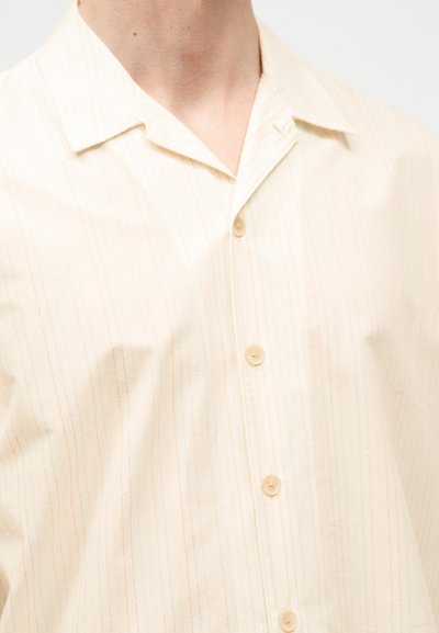 Κοντινό πλάνο ενός ατόμου που φοράει πουκάμισο σε κρεμ απόχρωση με κοντά μανίκια, κουμπιά και διακριτικές κάθετες ρίγες, με γιακά τύπου καμπ.