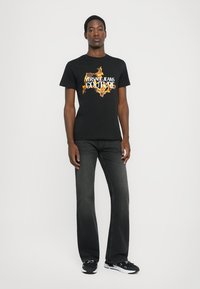 Črna bombažna majica s živahnim grafičnim dizajnom, ki vsebuje "Versace Jeans Couture" v beli in zlati barvi. Ujemajoča se temna kavbojka in teniske.
