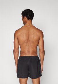 Schwarze Badeshorts mit elastischem Bund, leichtem Stoff und glatter Textur, von hinten betrachtet. Minimalistisches Design, keine sichtbaren Muster.