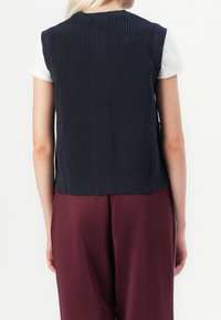 Tmavě námořnická pletená vesta s žebrovanou strukturou, bez rukávů a s rovnou spodní částí, kombinovaná s světlým topem a vínovými kalhotami.