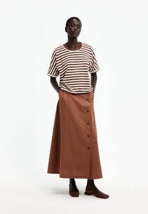 Frau trägt ein braun-weiß gestreiftes kurzärmeliges Hemd, einen braunen, geknöpften langen Rock und braune flache Schuhe, steht mit den Händen in den Taschen.