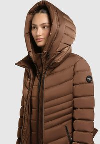 Braune Daunenjacke mit einer großen, gepolsterten Kapuze. Verfügt über ein horizontal gestepptes Muster, Reißverschlüsse und ein schwarzes Logo-Detail am Ärmel.