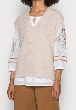 Femme portant un gilet sans manches beige en maille sur une blouse blanche avec broderies florales sur les manches et un pantalon marron sur fond blanc.