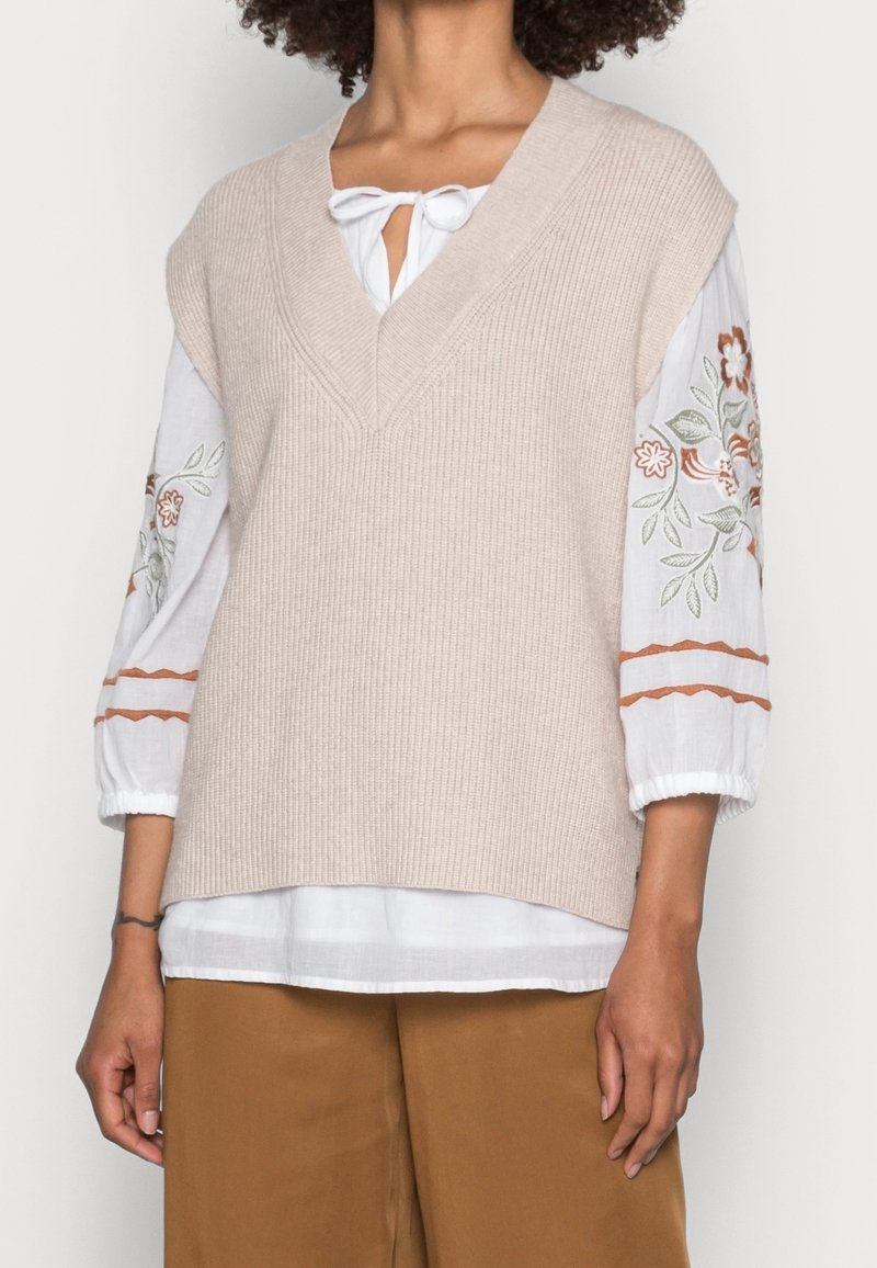 Femme portant un gilet sans manches beige en maille sur une blouse blanche avec broderies florales sur les manches et un pantalon marron sur fond blanc.