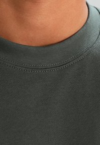Primo piano di una maglietta in cotone verde scuro, con collo rotondo a costine e texture liscia, caratterizzata da cuciture uniformi lungo il colletto.