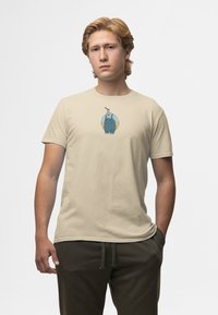 Beige katoenen T-shirt met een cartoonfiguurtje in blauwe overall, met een afgeronde vorm en een gladde textuur. Model draagt donkere broek.