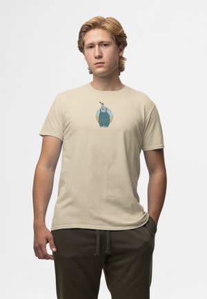 Beige bomullst-shirt med en tecknad figur i blå hängselbyxor, med en rundad form och en slät textur. Modellen bär mörka byxor.