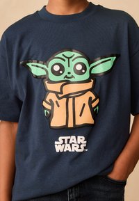 Navy blue cotton t-shirt featuring a large graphic of a cartoon character with green skin and black eyes, wearing an orange coat and a "Star Wars" logo.