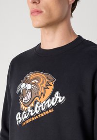 Fekete pulóver narancssárga és barna tigrist ábrázoló grafikával, fehér részletekkel. Az image alatt a "Barbour International" szöveg található.