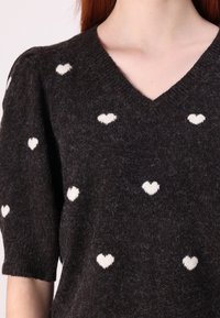 Maglione lavorato a maglia scura con scollo a V, maniche corte a sbuffo, caratterizzato da un motivo di cuori bianchi su tutto il capo. Tessuto morbido e vestibilità comoda.