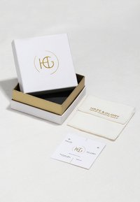 Boîte cadeau blanche avec des accents dorés, portant le logo "H&G". Comprend une pochette à bijoux crème et une étiquette blanche avec du texte doré. Texture lisse.