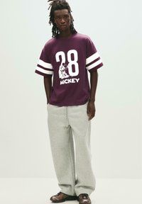 Paarse oversized T-shirt met witte strepen, met een grote "28" en Mickey Mouse grafiek; gecombineerd met lichtgrijze joggingsbroek en donkere sneakers.