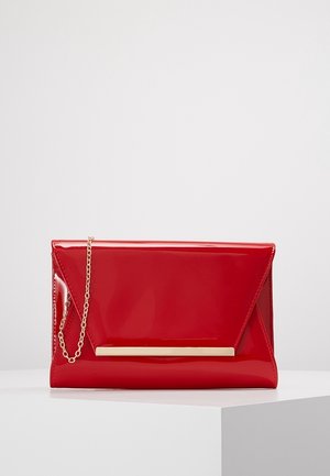 Rote Klappentasche aus Lackleder mit goldener Stabdetails und kleiner goldener Kettenriemen auf weißer Fläche vor hellgrauem Hintergrund.