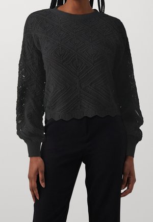 Vrouw draagt een zwarte gebreide trui met lange mouwen, een geometrisch patroon en golvende zoom, gecombineerd met een zwarte broek.