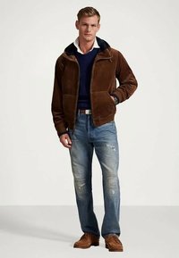 Καφέ φούτερ μπουφάν με φερμουάρ και μαύρη κουκούλα, μπλε πουλόβερ από κάτω, σκισμένα ανοιχτό μπλε τζιν και καφέ παπούτσια.