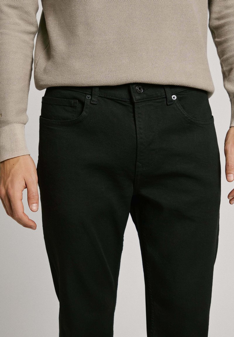 Schwarze Jeans mit slim fit, ausgestattet mit einem Knopfverschluss, fünf Taschen und einer subtilen Textur. Tragen mit einem beigen Strickpullover.