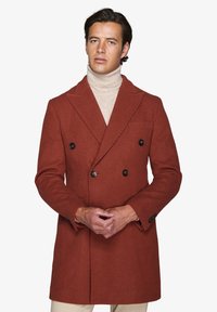 Dobbeltradet frakke i rustfarvet tekstureret stof med sorte knapper, udskårne klapper og sidelommer. Båret over en beige turtleneck.
