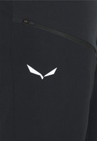Pantaloni atletici negri cu un buzunar cu fermoar și un logo alb cu forme unghiulare în partea de jos. Textură de țesătură fină, design aderent.