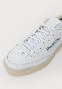 Λευκό δερμάτινο αθλητικό παπούτσι με διάτρητο κουτί δακτύλων, επίπεδα κορδόνια και διαφανές καουτσούκ σόλα. Διαθέτει πράσινο λογότυπο Reebok στο πλευρό.