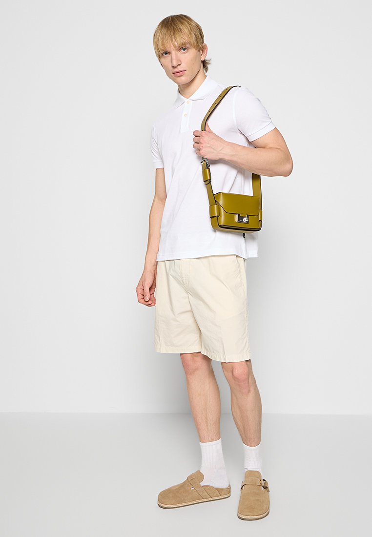 Chemise polo blanche, shorts beiges, chaussures slip-on tan. Tient un sac bandoulière jaune au design épuré et avec un matériel proéminent.