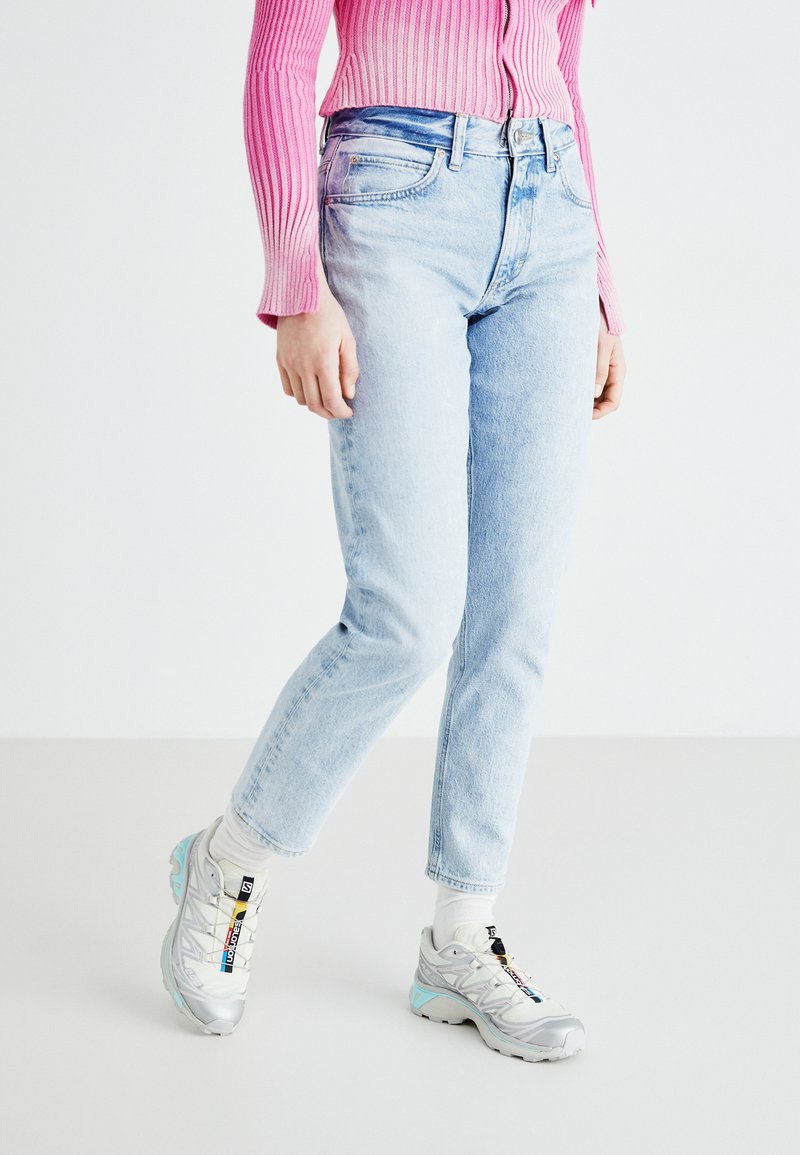 Vaqueros de mezclilla azul claro con un corte recto, combinados con un suéter de rayas rosa. Llevados con zapatillas de correr en gris y verde azulado.